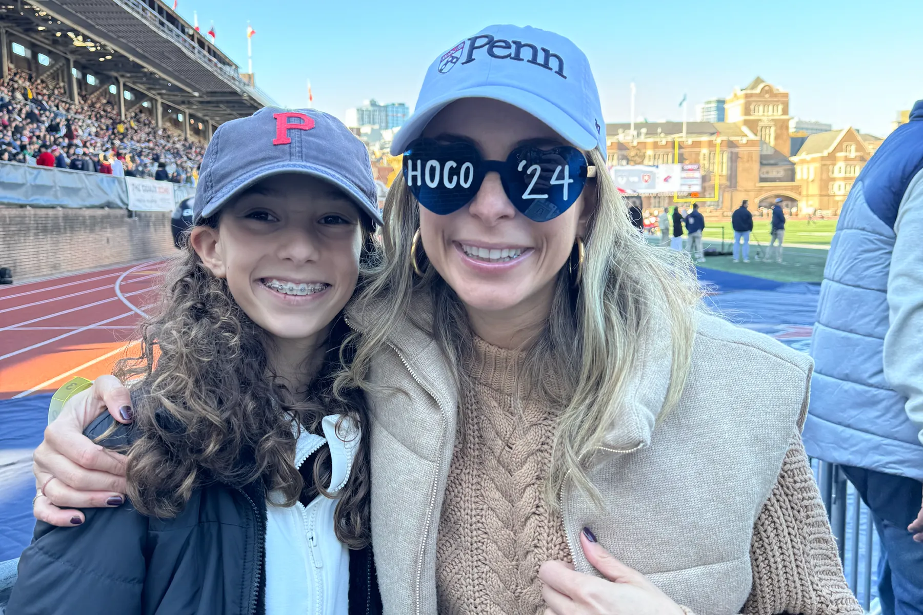 Two people dressed in casual fall outfits and Ivy League hats smiling for a photo at a busy outdoor stadium during a homecoming celebration.