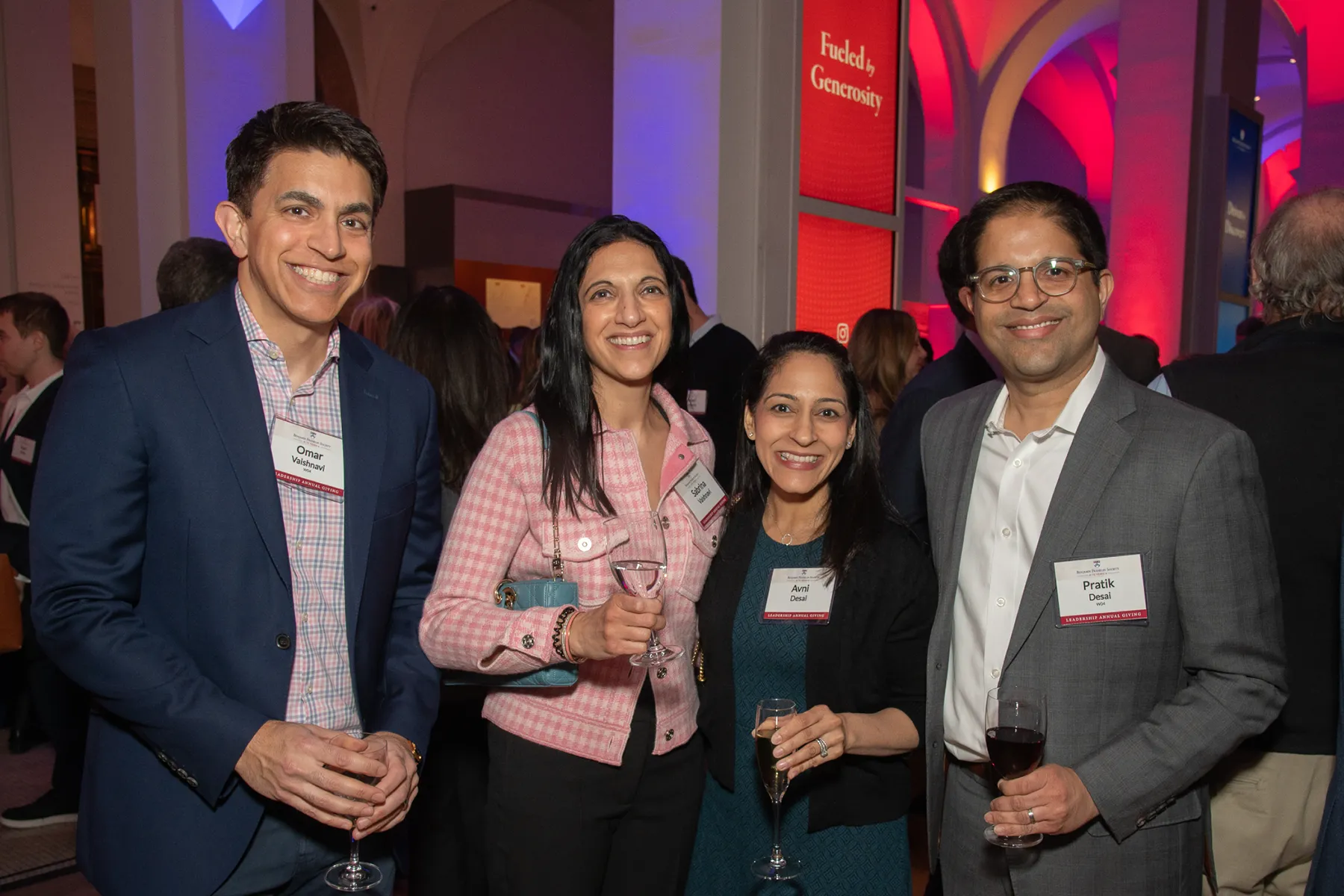Four guests pose together at a University of Pennsylvania evening event, smiling and holding drinks in a warmly lit reception with red and blue lighting.