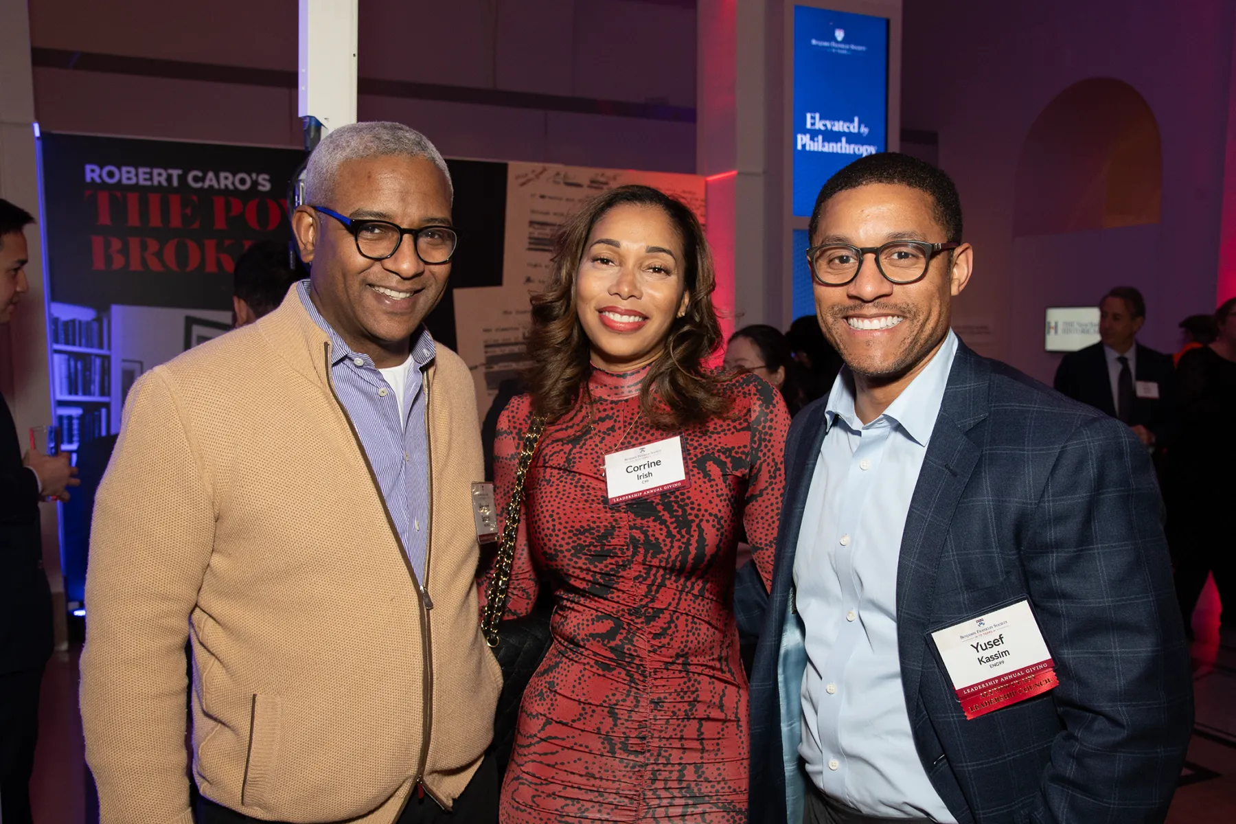 Three Penn alumni smile together at an evening celebration, dressed in formal-casual attire and wearing name tags in a venue lit with blue and red lights.