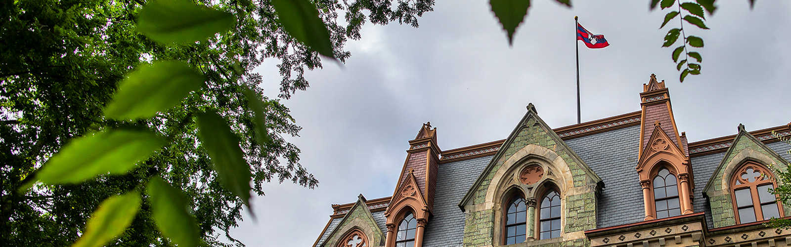 The top of College Hall at the University of Pennsylvania, with the University flag waving above the building and lush foliage in the foreground.