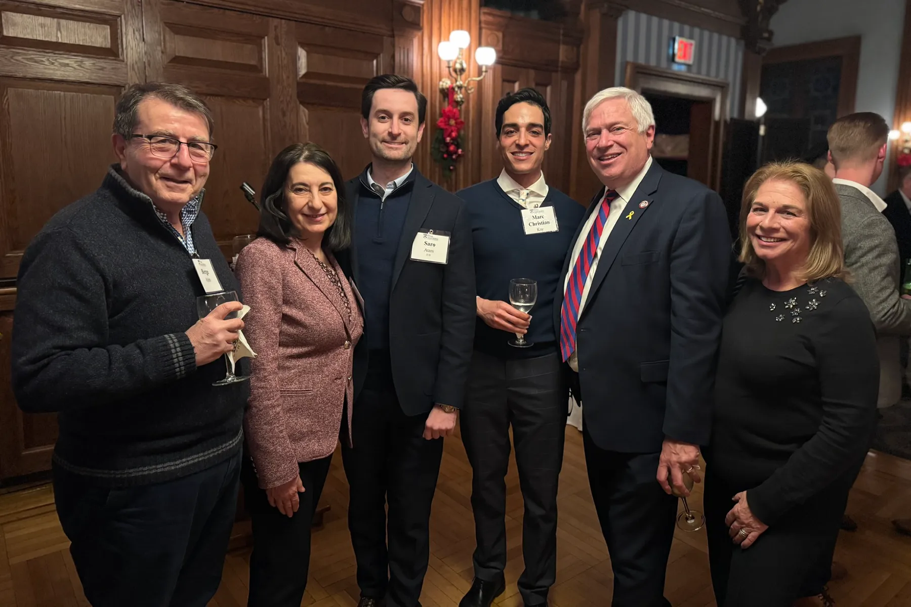Six people pose together at a University of Pennsylvania alumni event, dressed in semi-formal attire and holding drinks in a warmly lit wood-paneled room.