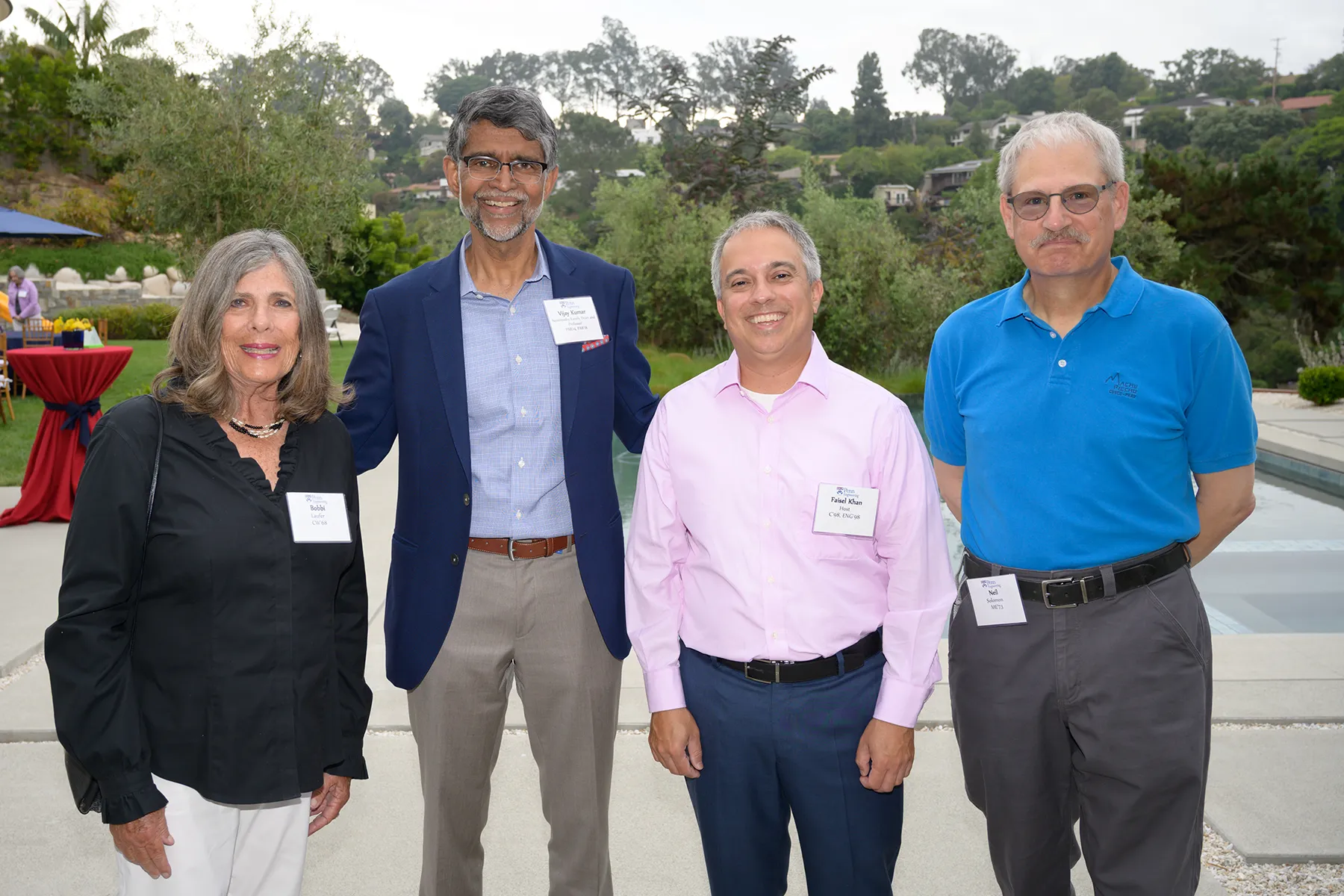 Four guests stand together at an outdoor Penn alumni event near a pool, smiling and wearing name tags with greenery and hills in the background.