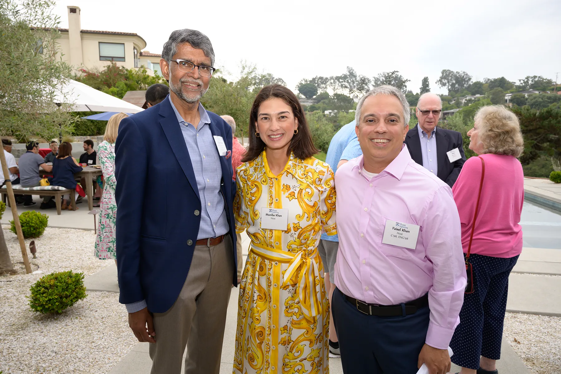 Three smiling guests pose outdoors at a University of Pennsylvania alumni event, with a pool, trees, and seated attendees in the background.