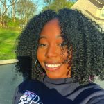 Selfie of a joyful young woman outdoors, wearing a 'University of Pennsylvania' sweatshirt. She is smiling brightly, with a suburban background featuring trees and a house, all under clear sunny conditions.
