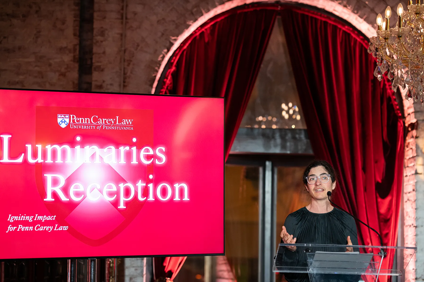 A speaker at the podium addresses the audience at the Penn Carey Law Luminaries Reception, with a large event screen and red drapery in the background.
