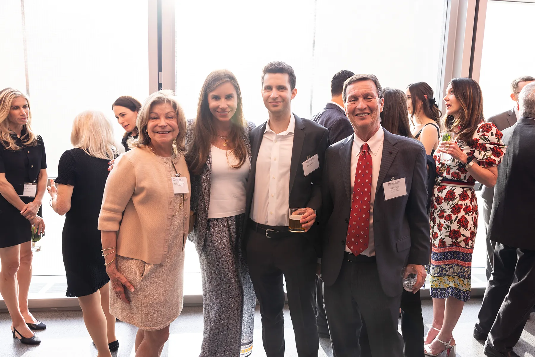 A group of four guests poses for a photo at a University of Pennsylvania event held in a bright, sunlit space filled with attendees in business attire.