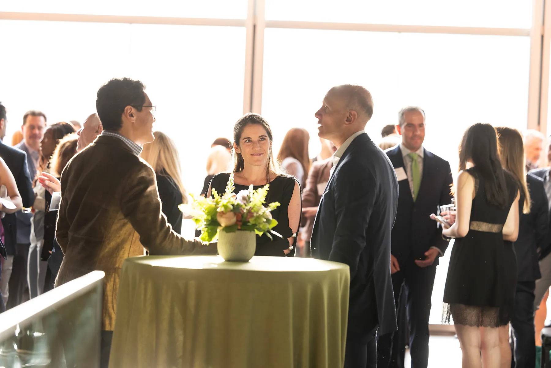 Three guests stand around a table with a flower arrangement, engaged in conversation during a Penn reception in a sunlit venue.