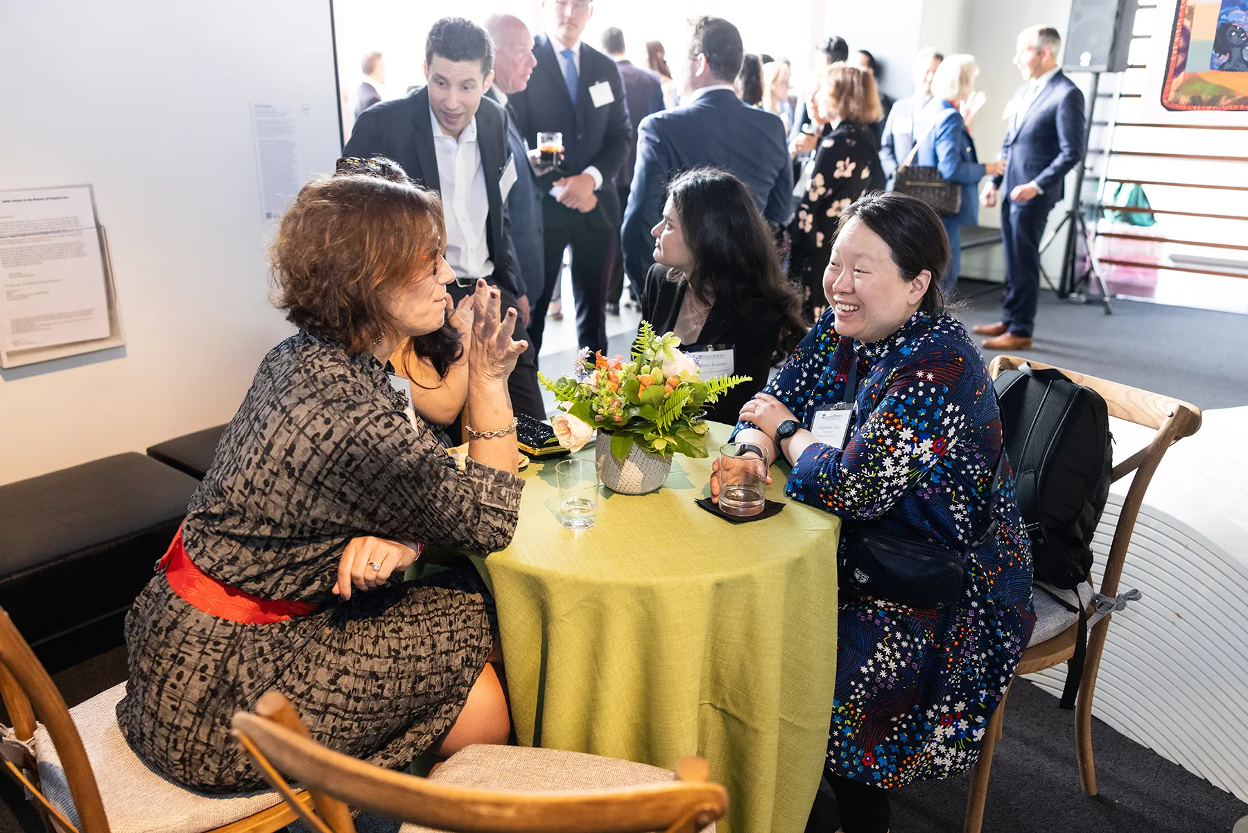 Guests enjoy conversation around a small round table with a green tablecloth and floral centerpiece during a Penn alumni reception in a modern indoor space.