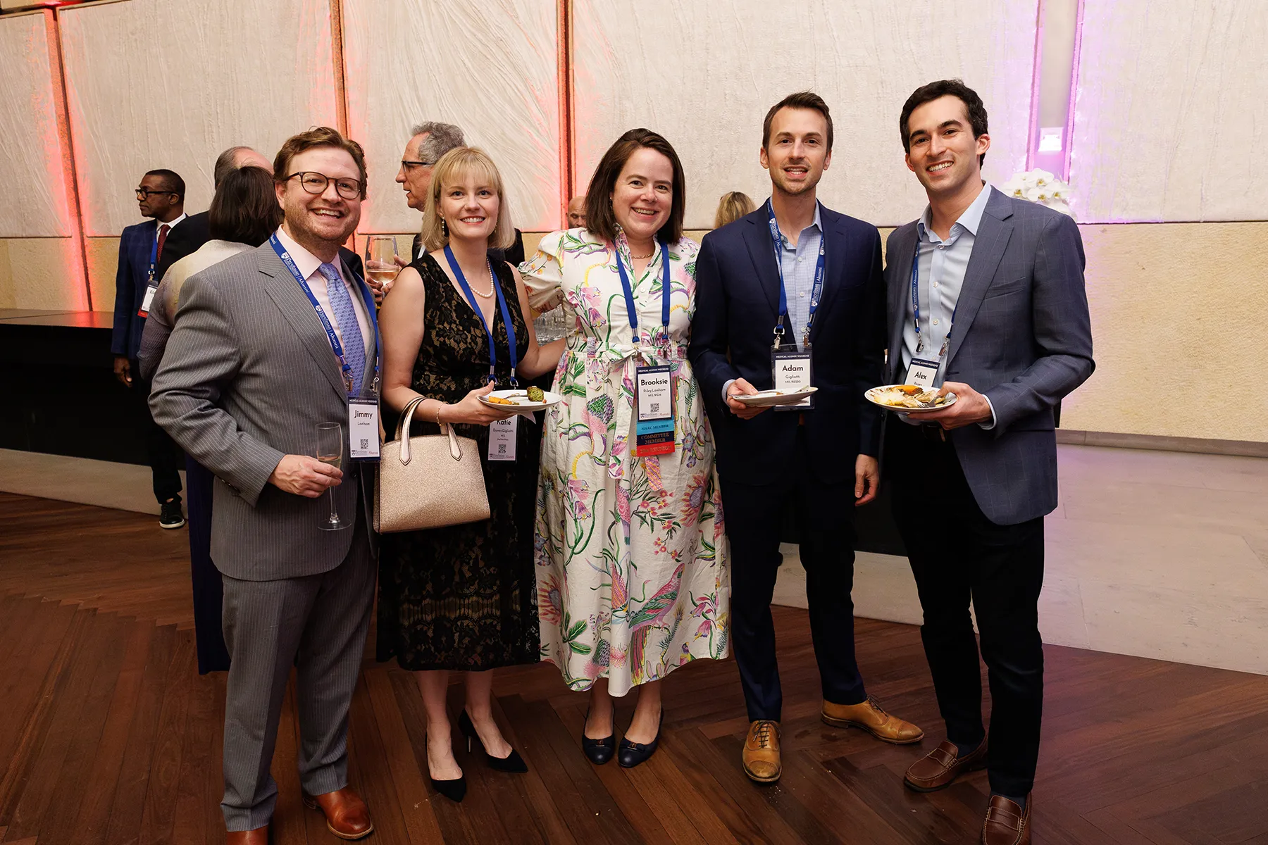 A group of five guests at a Penn alumni event pose together, holding plates and drinks, wearing name badges and lanyards in a warmly lit reception space.
