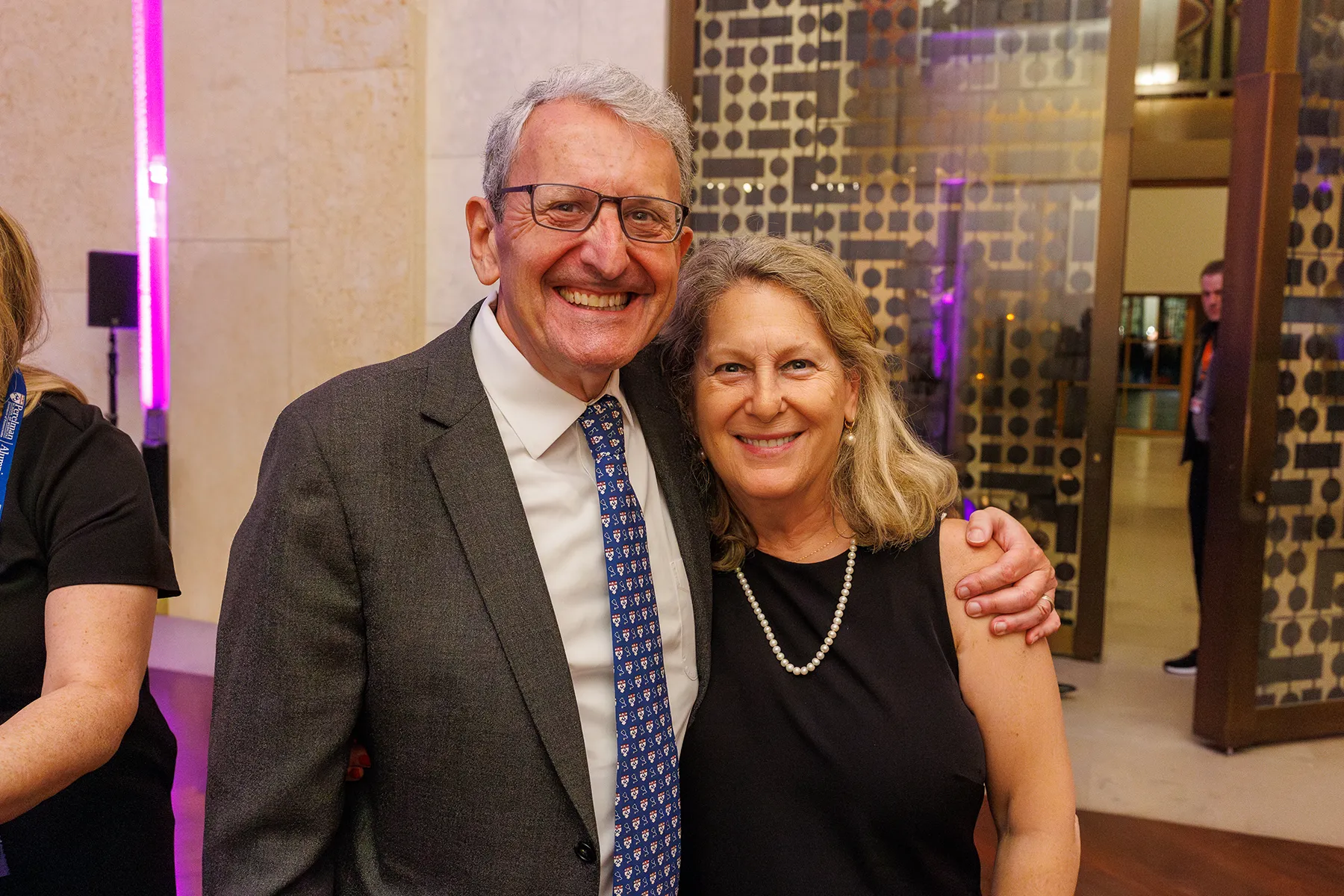 A man in a suit and a woman in a black dress pose together, smiling at a Penn event held in an elegant venue with soft lighting and patterned glass doors.