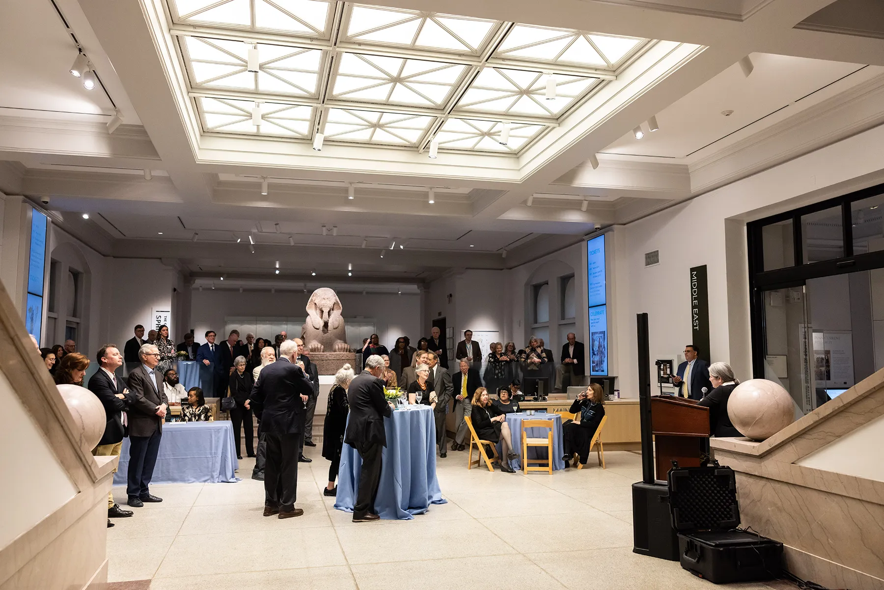 A crowd attends a University of Pennsylvania reception in the Penn Museum, seated and standing around cocktail tables with a central sphinx statue in view.