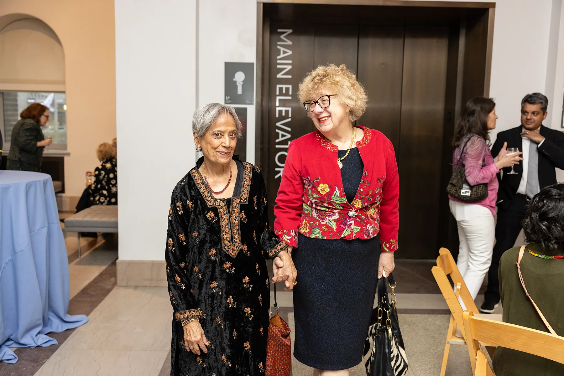 Smiling attendees hold hands while walking past the main elevator at the Penn Museum during a University of Pennsylvania reception.