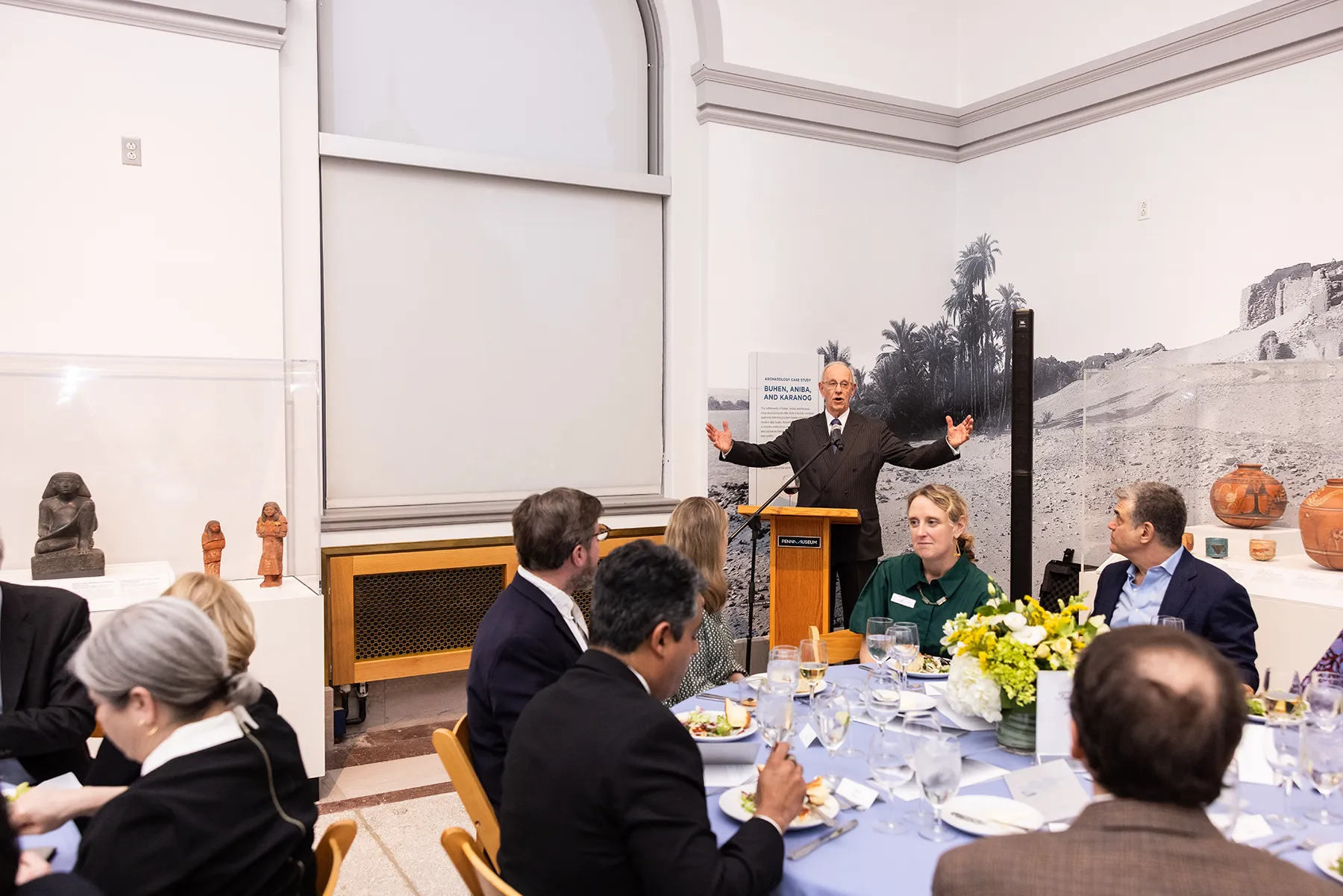A speaker gestures from a podium during a formal dinner at the Penn Museum, surrounded by guests seated at round tables with flowers and table settings.