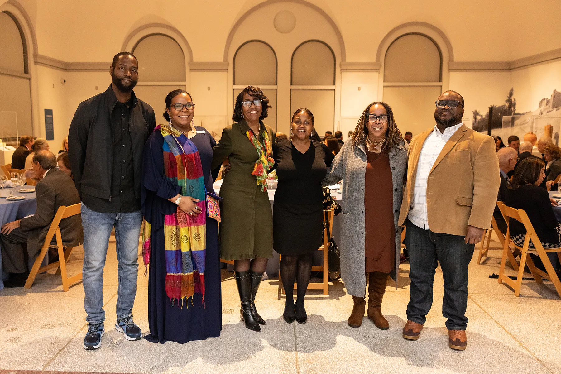 Six guests pose together at a Penn Museum dinner event, standing in a row and smiling in front of seated attendees and arched architectural details.