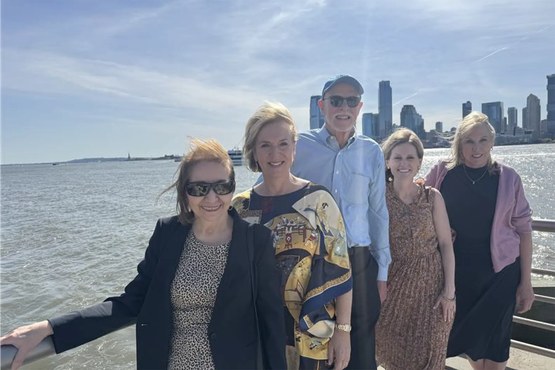 Five people stand together on a sunny waterfront promenade with the New York City skyline in the background and the Statue of Liberty visible in the distance.