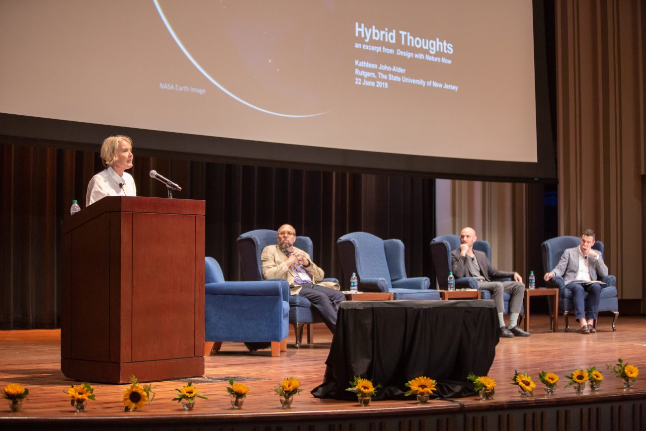 a woman at a podium on stage with five blue chairs and three of them have men sitting in them listening to the woman's speech