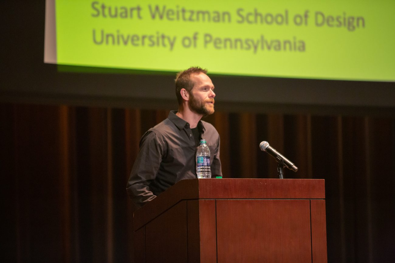 man standing at a podium giving a speech with a sign in the background saying stuart weitzman school of design university of pennsylvania