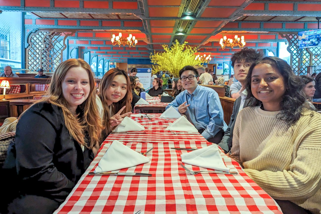 Five students sit together at a long table in a restaurant and smile for the camera
