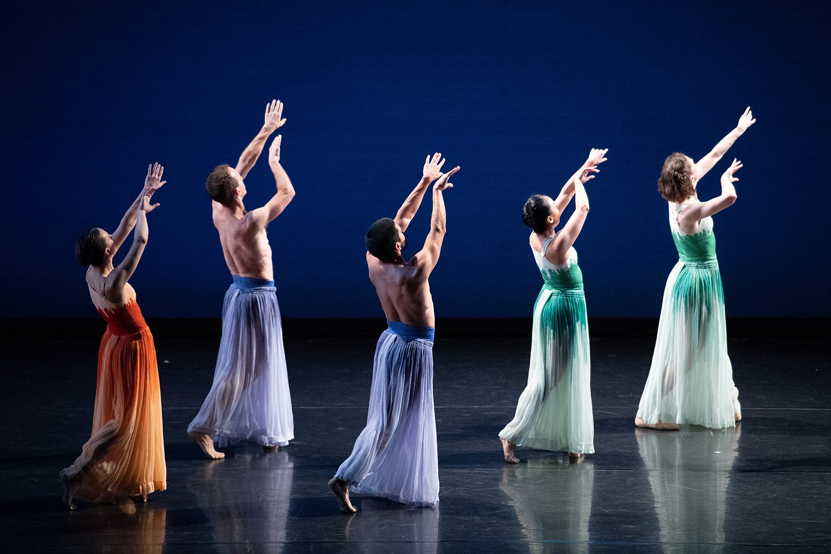 Five contemporary ballet dancers perform on a dimly lit stage, reaching gracefully upward in unison.
