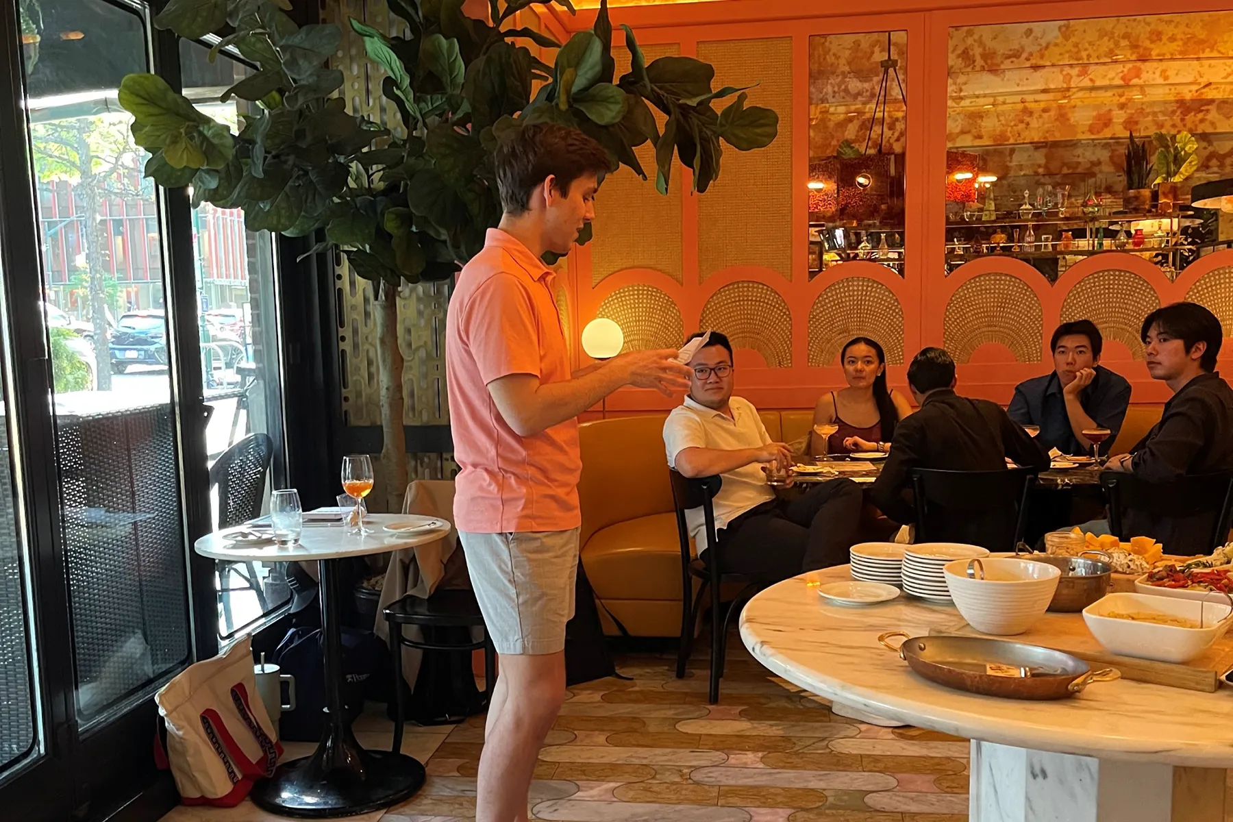 A man in a pink polo shirt speaks to a group of people seated at a restaurant table, with food and drinks on display in a warmly lit, colorful interior.