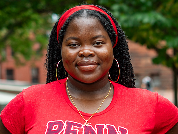 Herinah Asaah, C'26, smiling outdoors in a red Penn shirt on the University of Pennsylvania campus.