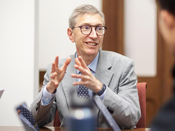 Russell Composto gestures while speaking in a meeting, seated at a conference table with laptops and colleagues.