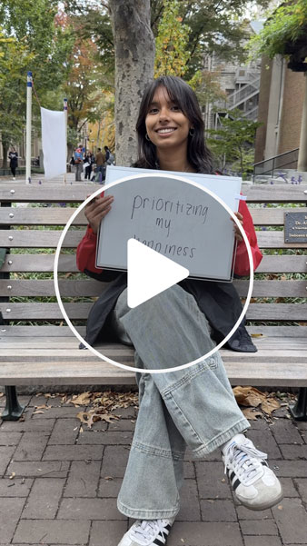 Student holding a whiteboard reading “prioritizing my happiness” on a campus bench.