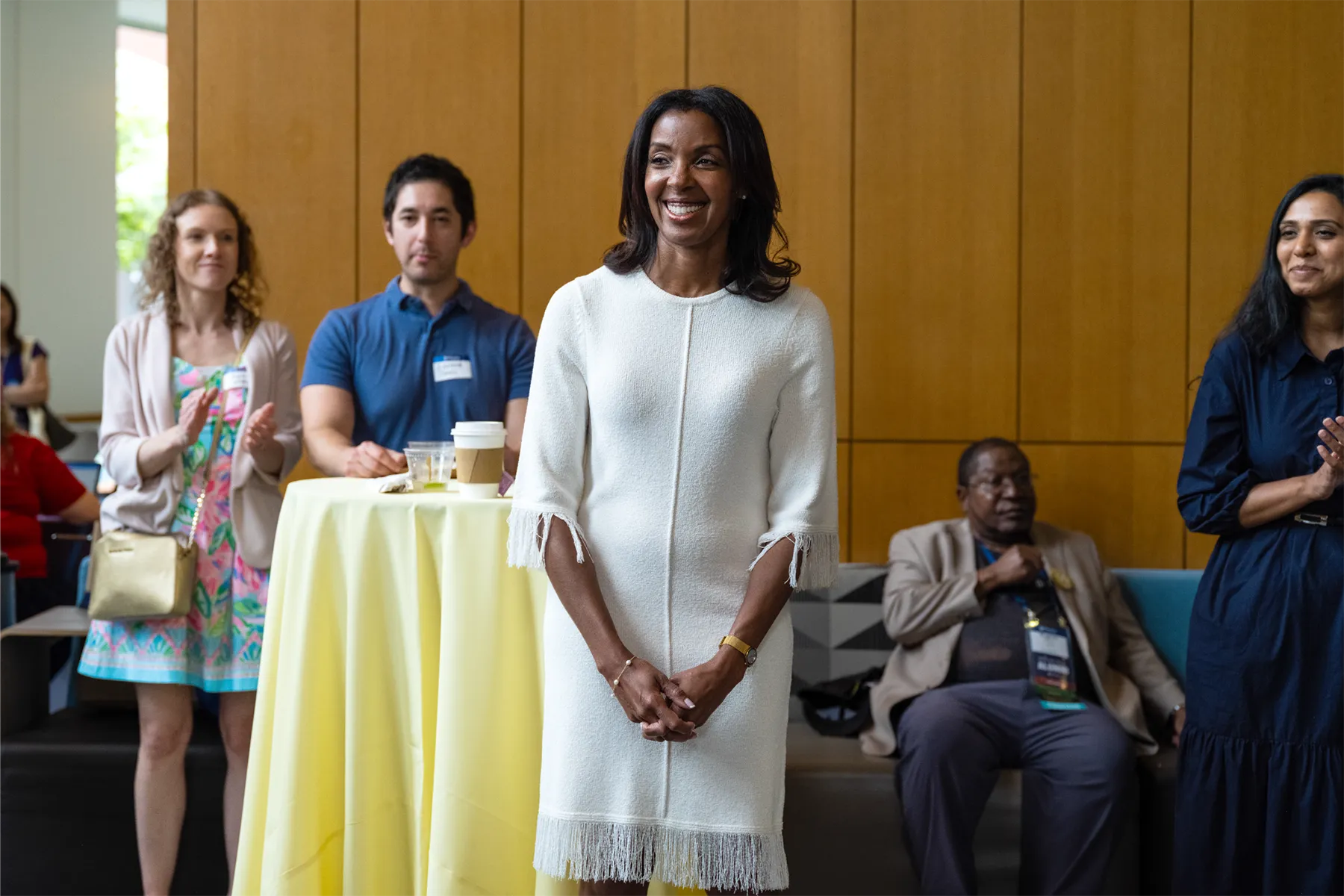A woman in a white dress smiles while standing in front of a group of applauding attendees during a University of Pennsylvania indoor event.