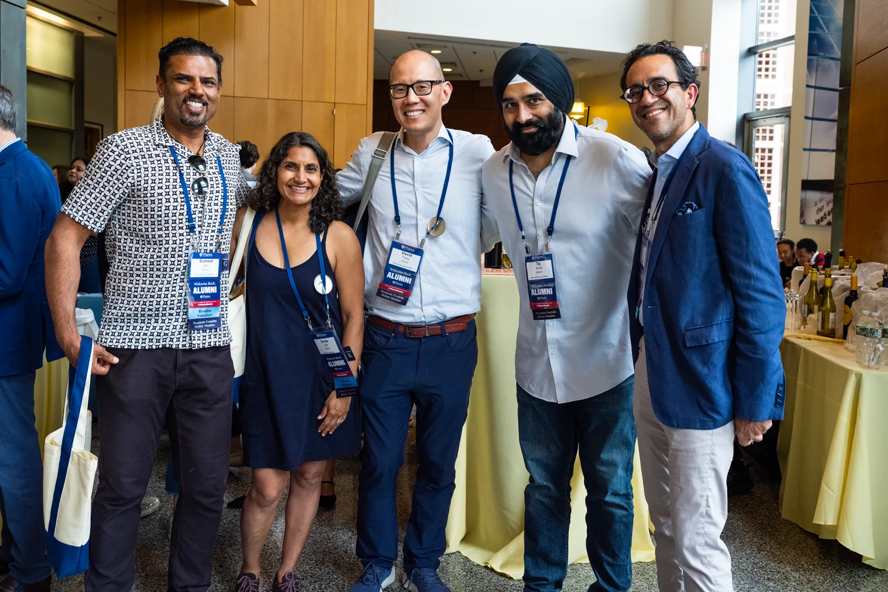 Five smiling attendees pose together at a University of Pennsylvania alumni event, wearing name badges and lanyards in a brightly lit indoor space.