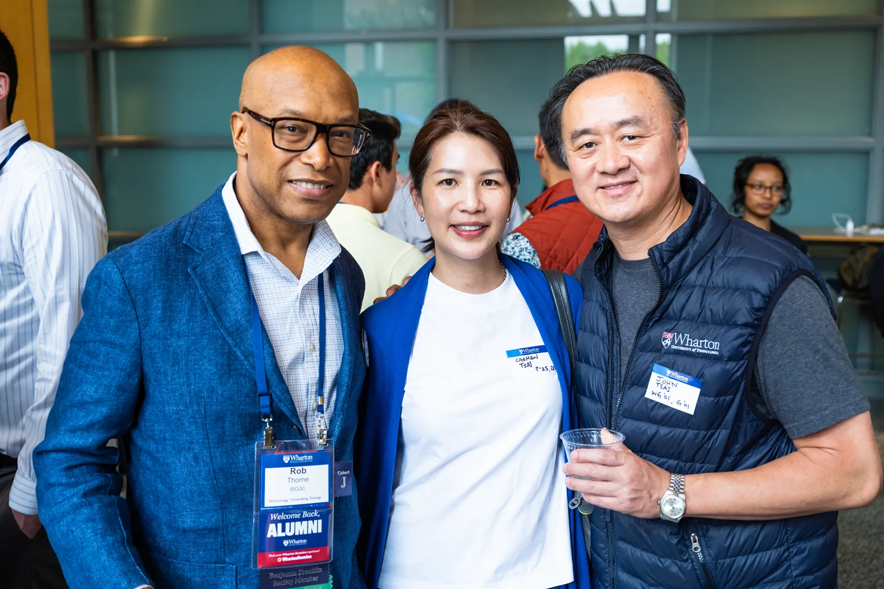 Three Wharton alumni smile together at a University of Pennsylvania event, wearing name tags and alumni badges in a modern indoor venue.