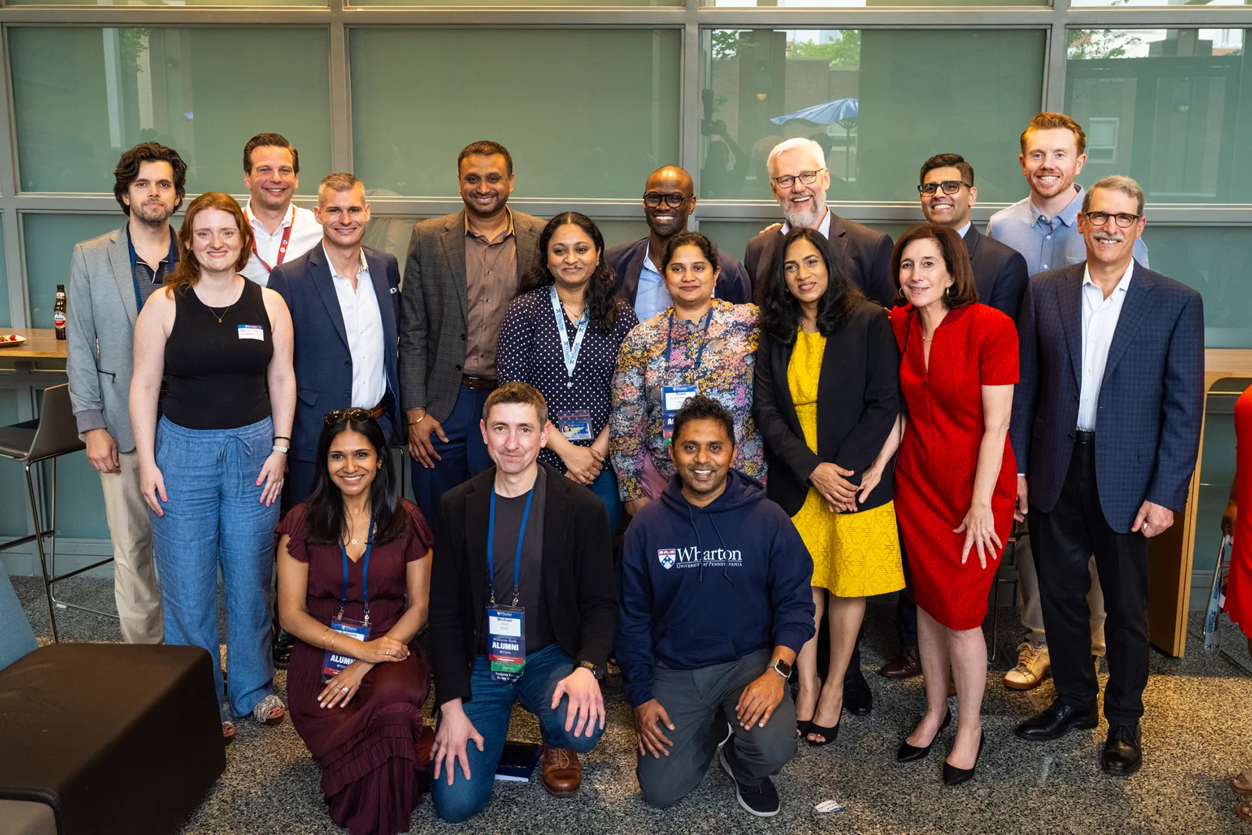 A large group of Wharton alumni poses together indoors during a University of Pennsylvania event, smiling and wearing badges in front of a glass wall.