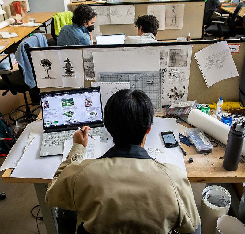 A graduate student in the Weitzman School of Design works at a busy workstation amid numerous fellow students