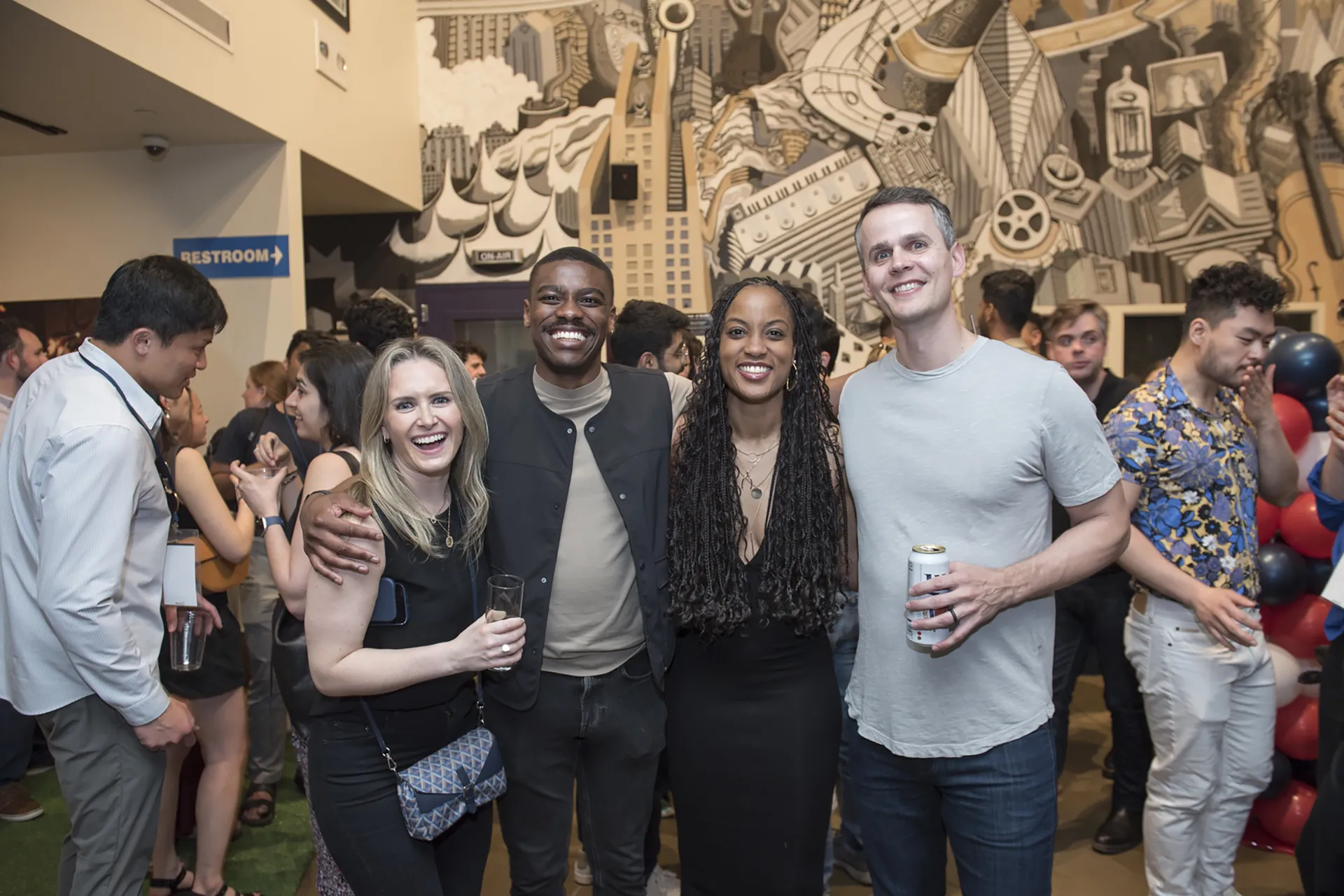 Four alumni stand arm-in-arm smiling in front of a large black-and-white mural at an Alumni Weekend indoor reception.