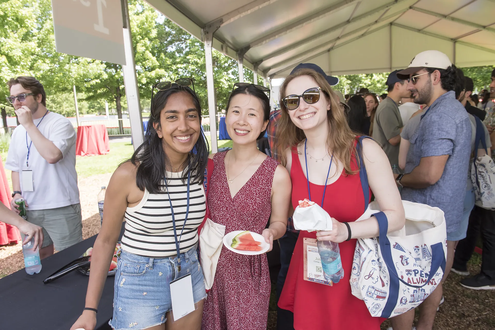 Trio of recent Penn graduates pose under a canopy, displaying event badges, tote bags, and summer snacks at Alumni Weekend.
