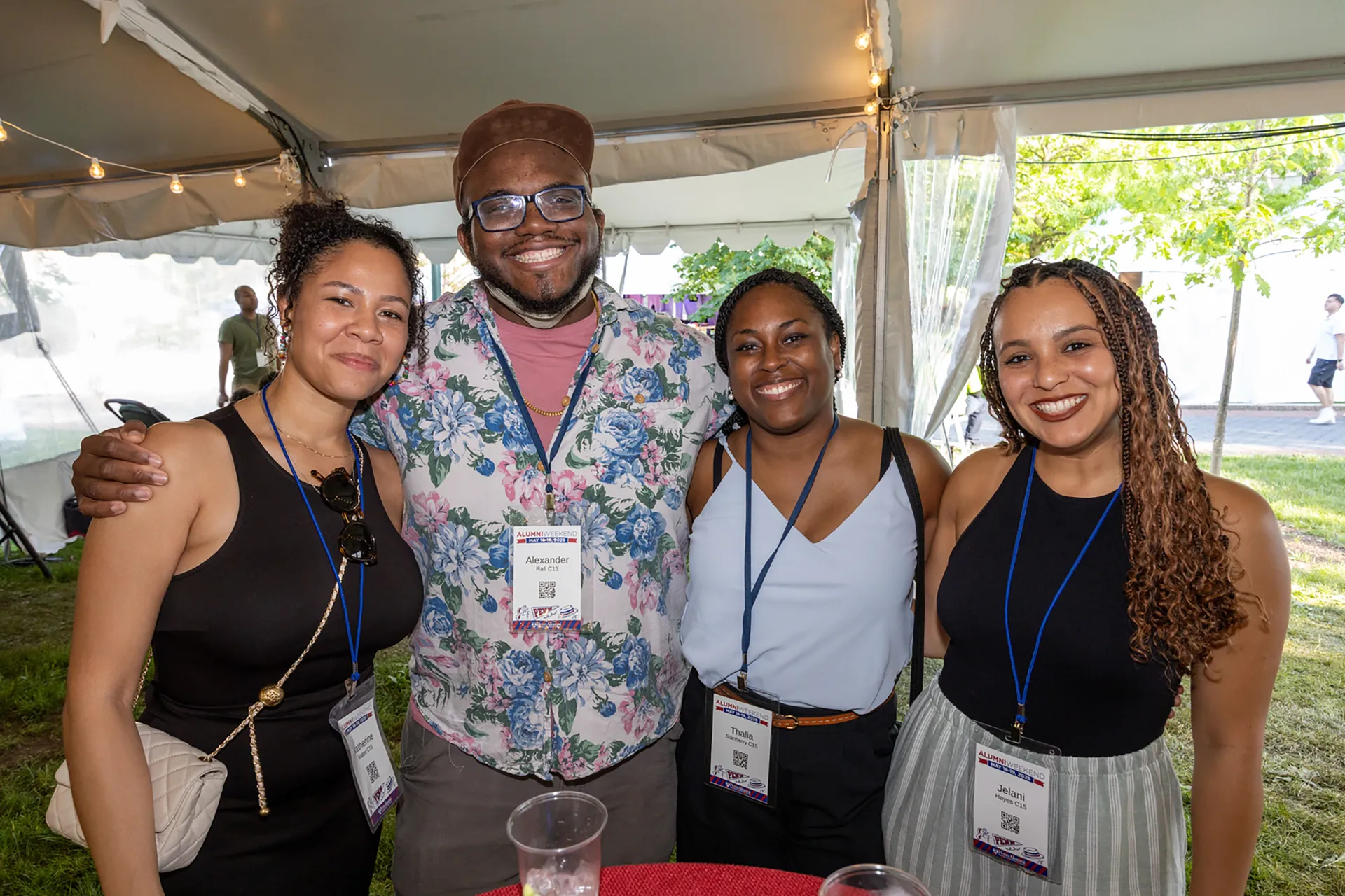 Four Penn alumni stand arm–in–arm under a white tent at Alumni Weekend, wearing name badges and smiling at the camera.