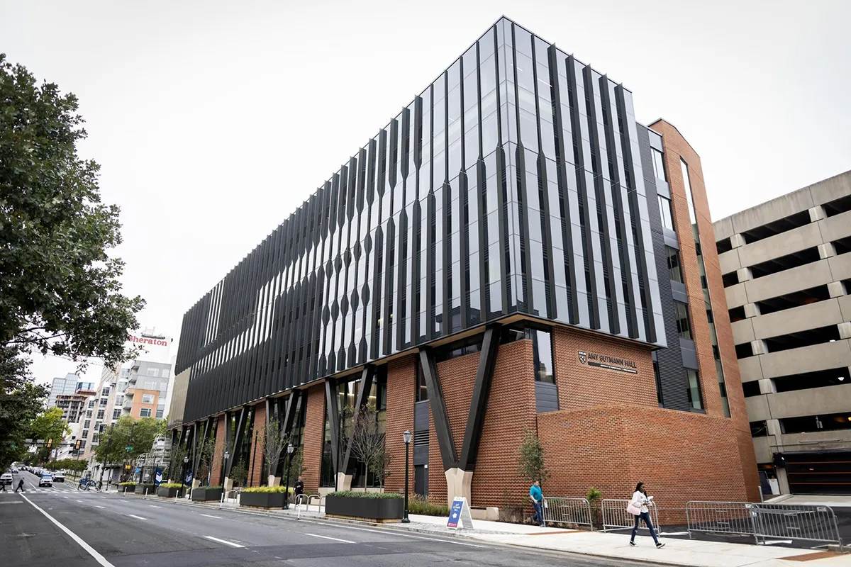 An exterior view of Amy Gutmann Hall, a modern, multi-story building with a striking architectural design featuring glass panels and black vertical slats on the upper facade, supported by angled steel beams and a brick base.