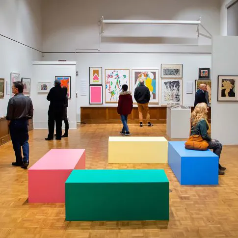 An exhibition of art at the Arthur Ross Gallery with people scattered throughout, looking at the items on display.