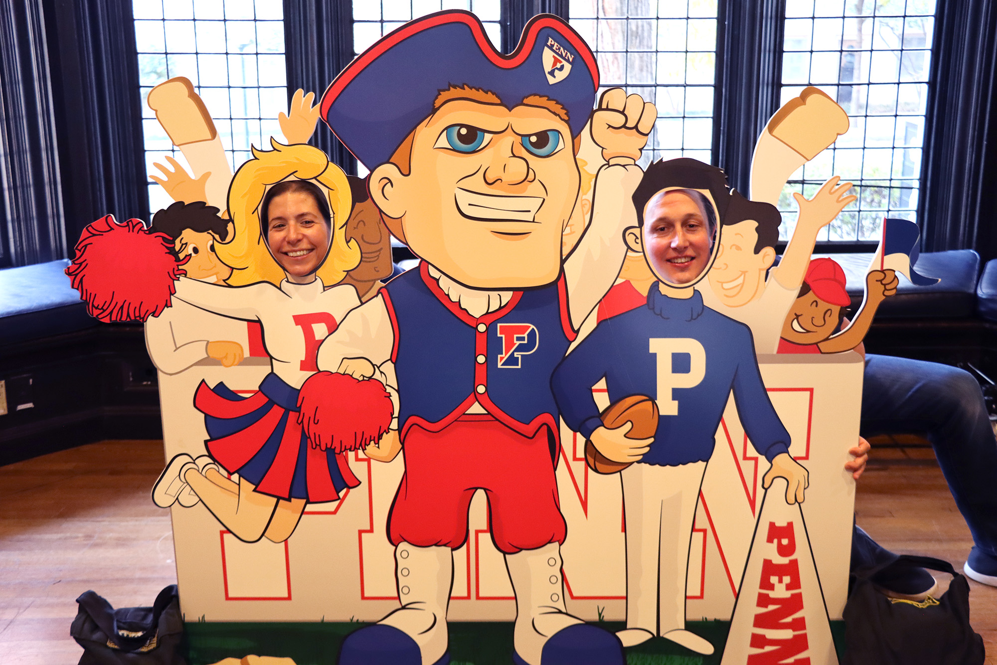 Two members of the Benjamin Franklin Society pose for a photo from behind a cutout of the Penn Quaker mascot during the 2022 Homecoming Brunch at the University of Pennsylvania.