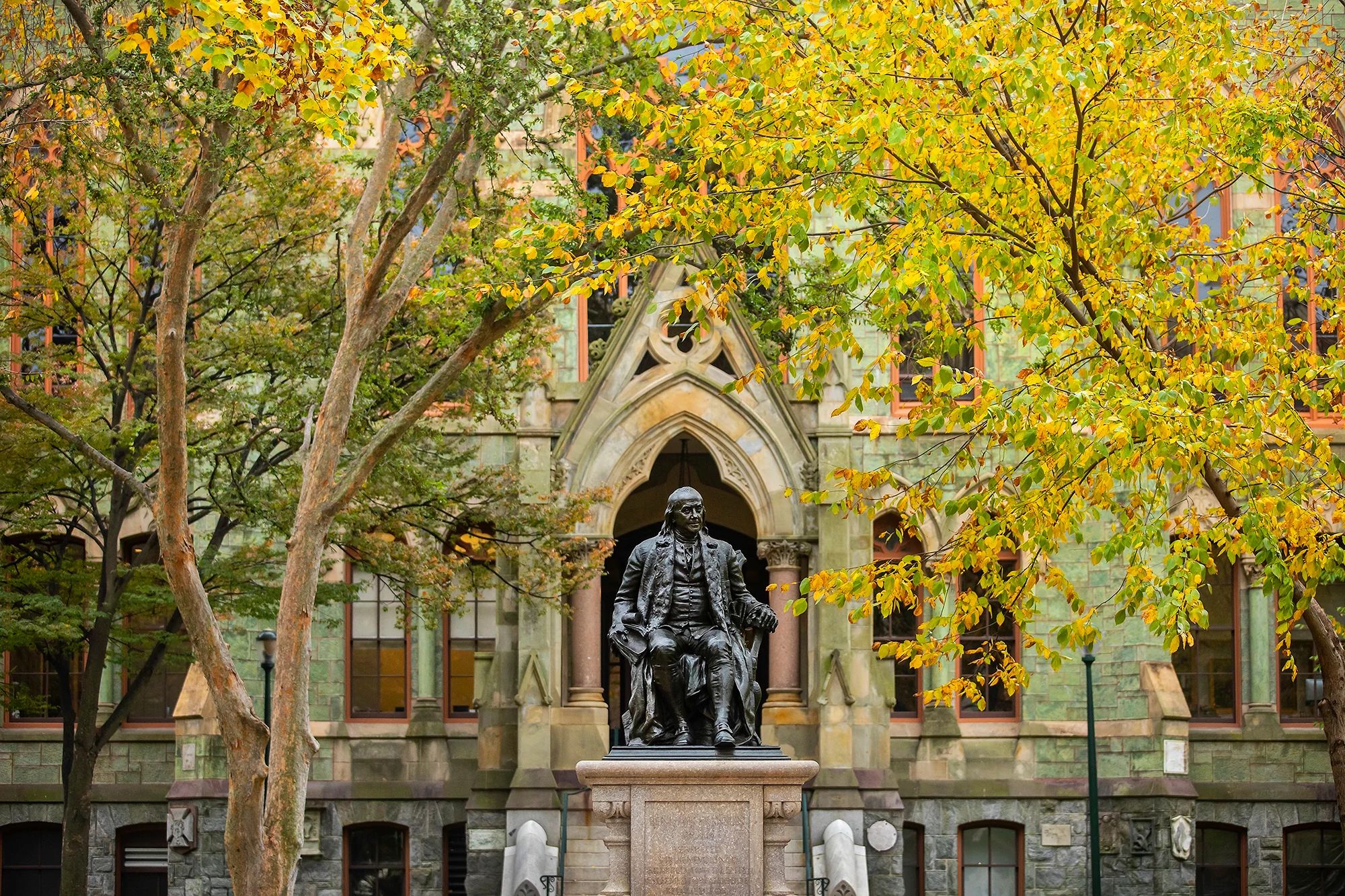 Statue of Benjamin Franklin seated in front of College Hall on College Green at the University of Pennsylvania, shown beneath changing leaves during fall