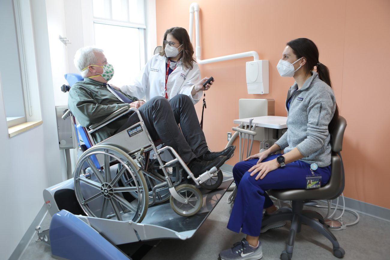 A patient in a wheelchair uses a chairlift while speaking to dental professionals during an appointment.