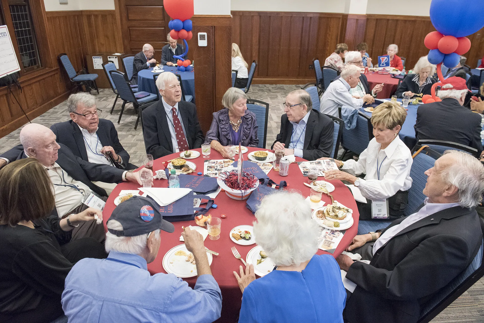 University of Pennsylvania Class of 1965 alumni seated around a red-clothed table enjoying food and conversation at their 60th reunion with red and blue balloon centerpieces
