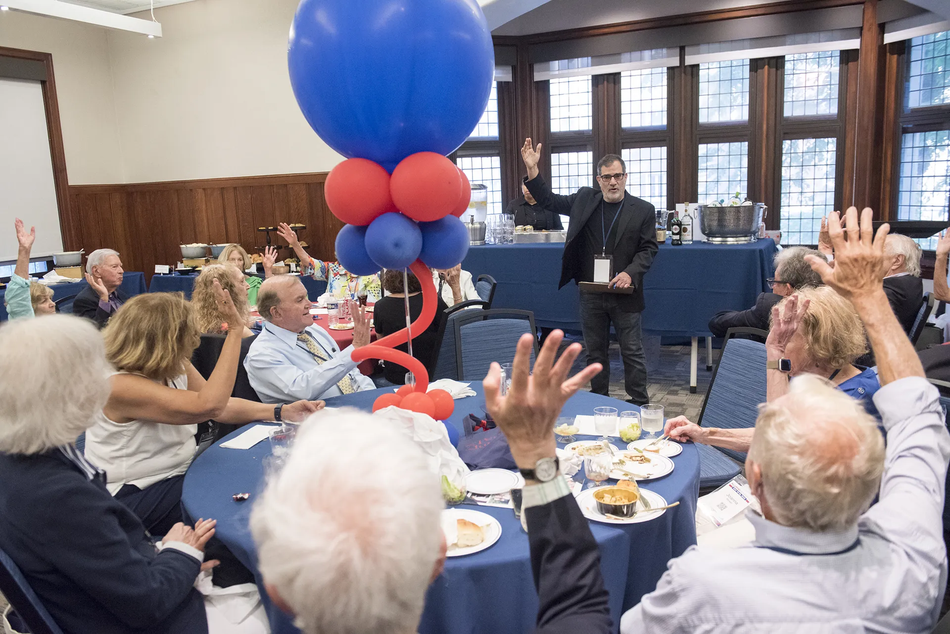 Penn ’65 reunion attendees interact with a speaker at the front of the room, all seated around tables decorated with Penn tote bags and patriotic balloons