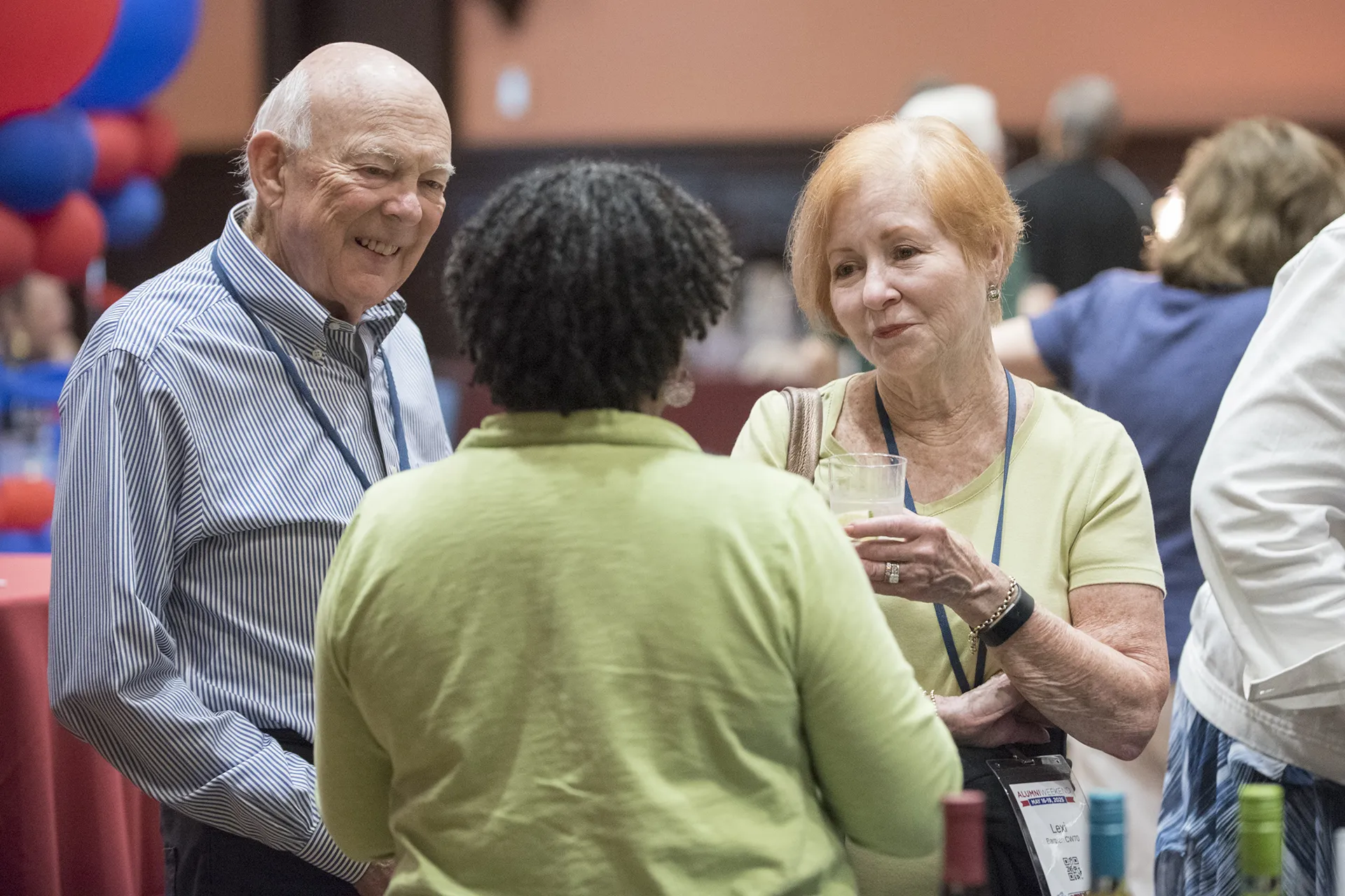 Three University of Pennsylvania Class of 1970 alumni chatting over drinks at their 55th reunion with red and blue balloon décor in the background