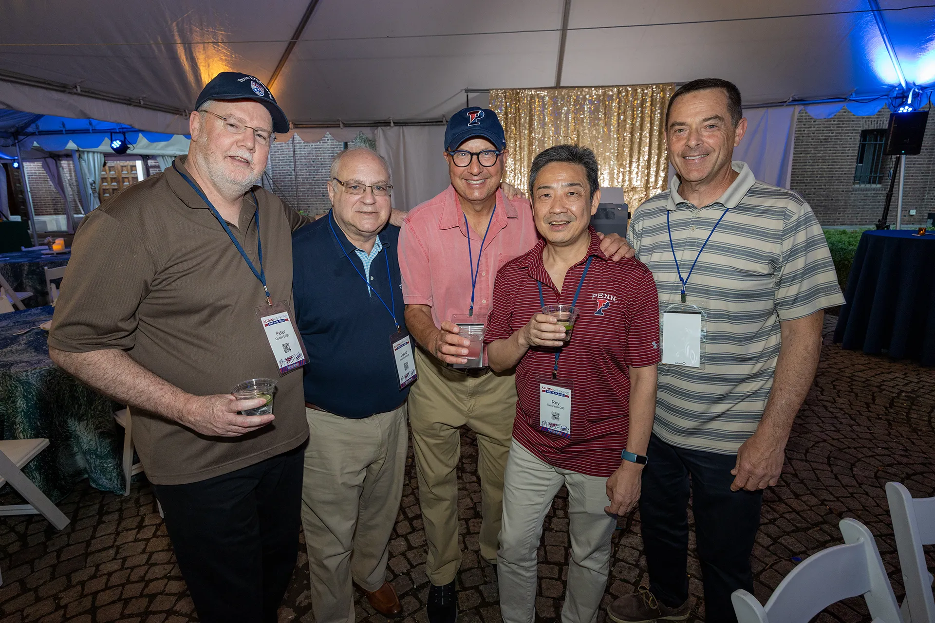 Five members of Penn’s Class of 1985 gathered under a white tent at their 40th reunion, wearing alumni lanyards and holding cocktails on a cobblestone patio