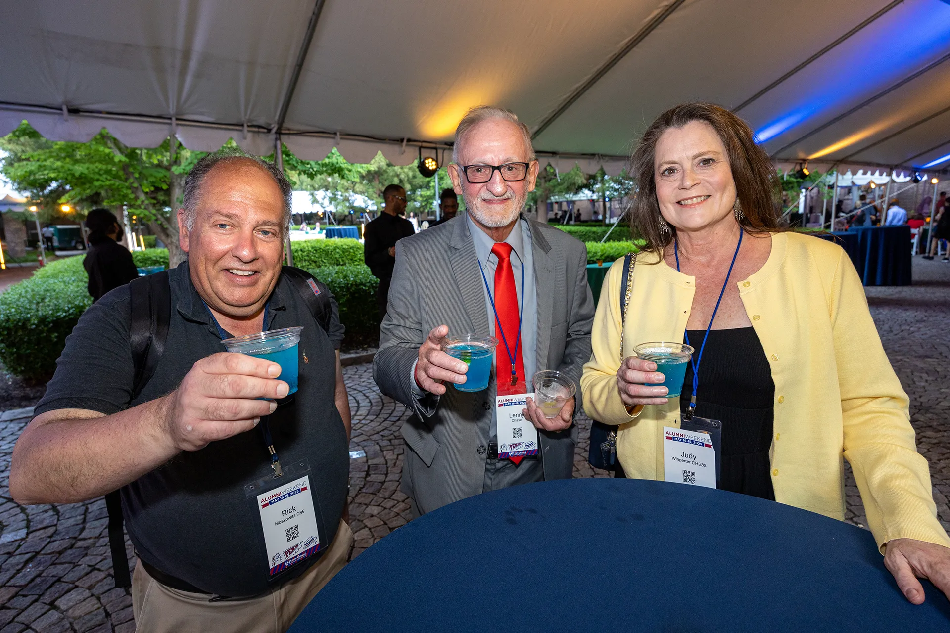 Three University of Pennsylvania Class of 1985 alumni raising blue cocktails under a white tent at their 40th reunion reception on a cobblestone courtyard