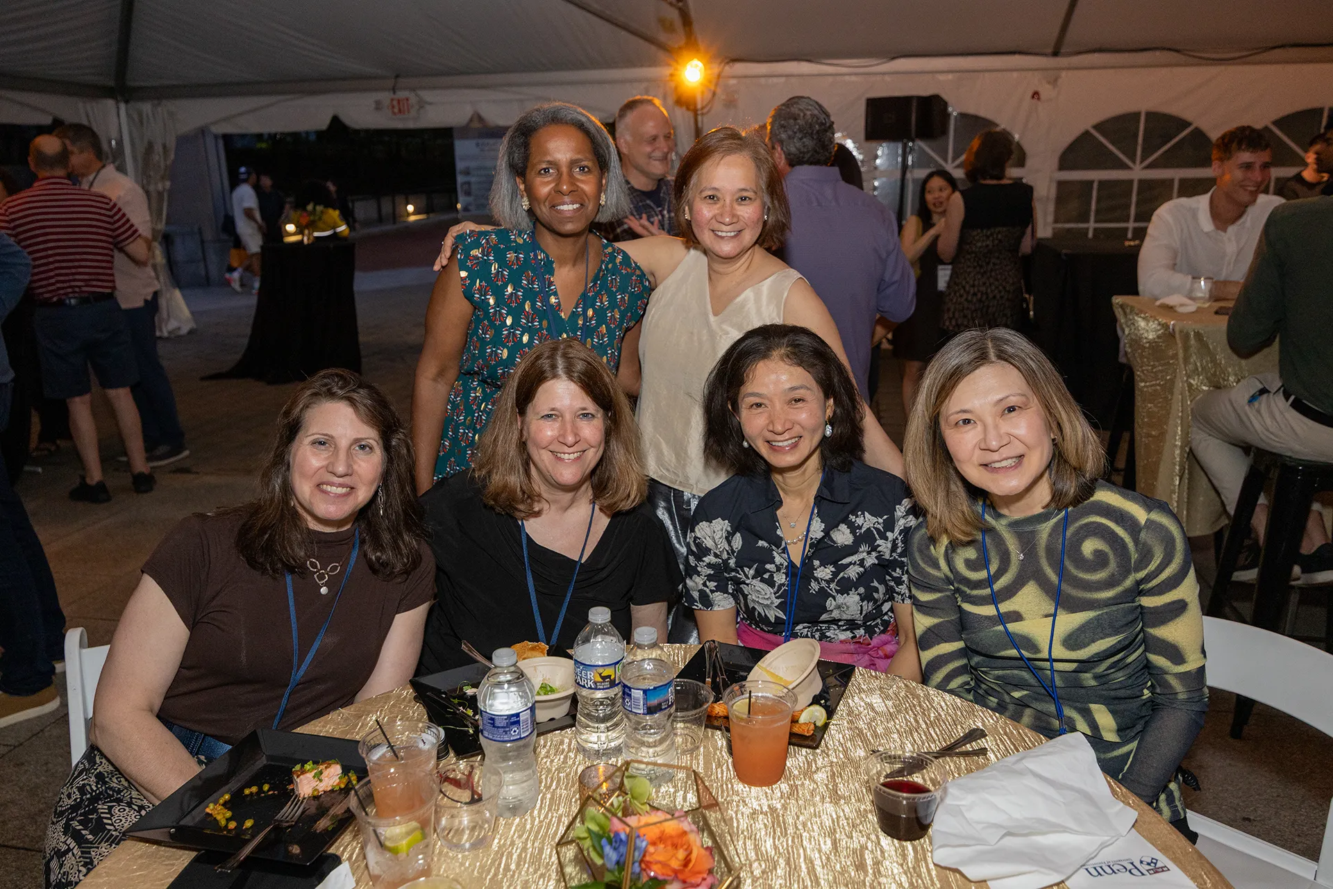 Six University of Pennsylvania Class of 1990 alumni—four seated around a table with reunion meals and drinks, two standing behind—smiling under a tent at their 35th reunion