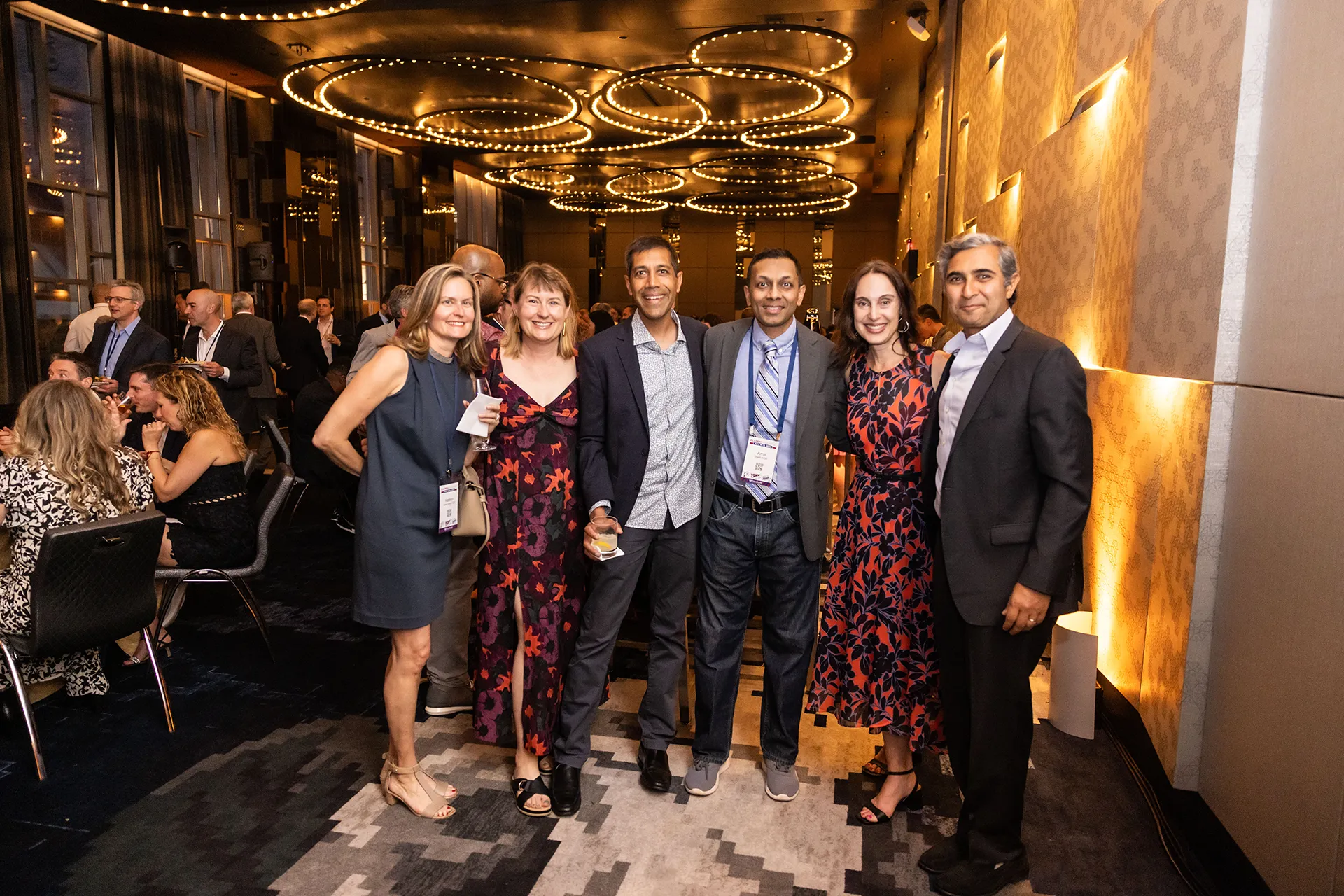 Six University of Pennsylvania alumni from the Class of 2000 standing arm-in-arm in a hotel lounge lit by circular ceiling lights during their 25th Reunion celebration