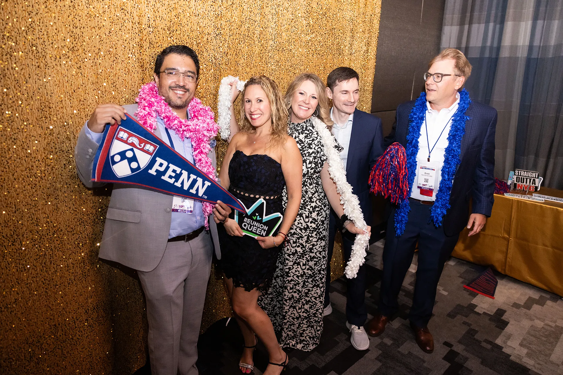 Five University of Pennsylvania alumni from the Class of 2000 posing with Penn pennant and festive boas against a gold sequin backdrop at their 25th Reunion photo booth