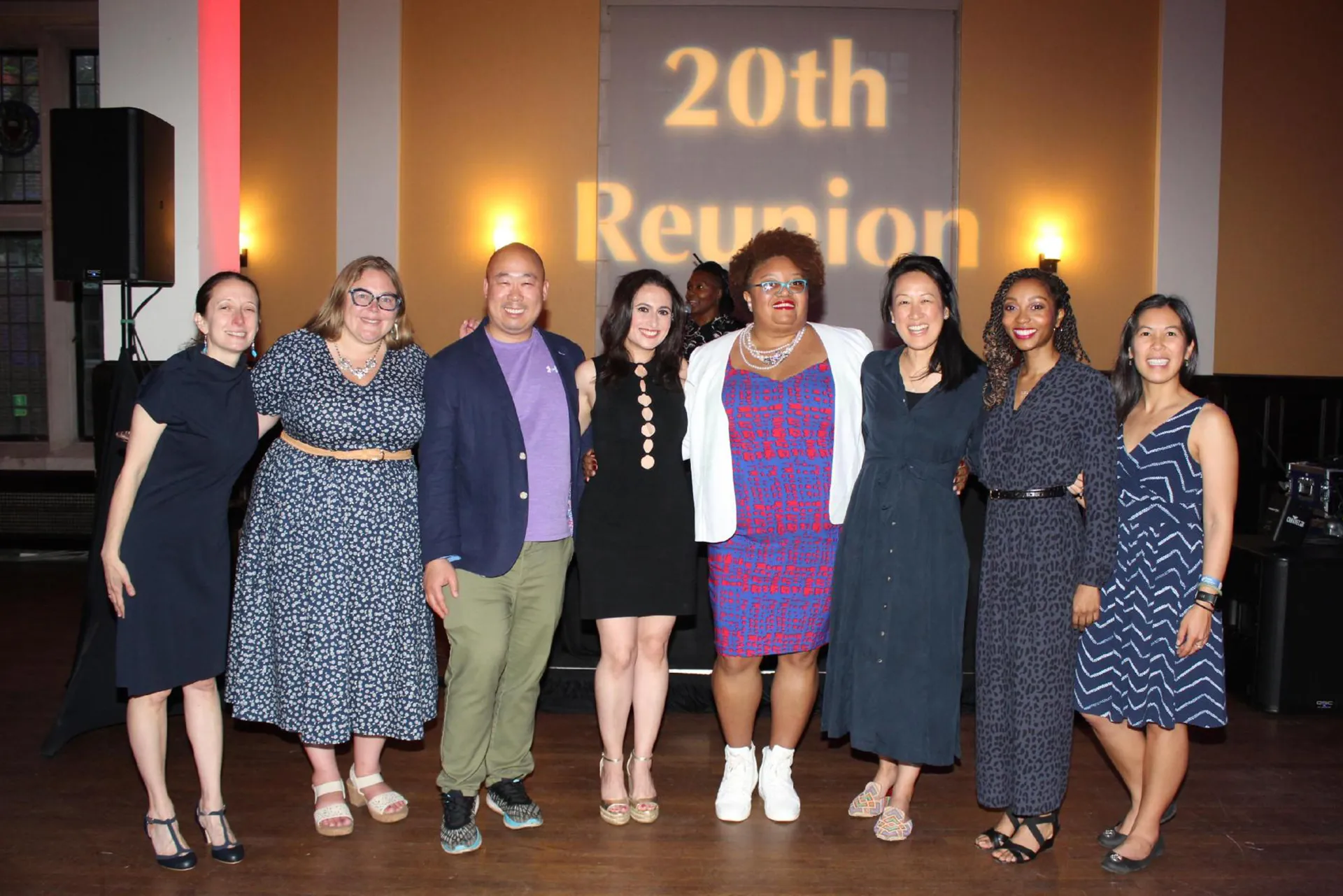 Eight University of Pennsylvania alumni from the Class of 2005 standing arm-in-arm under a “20th Reunion” projection in a reception hall
