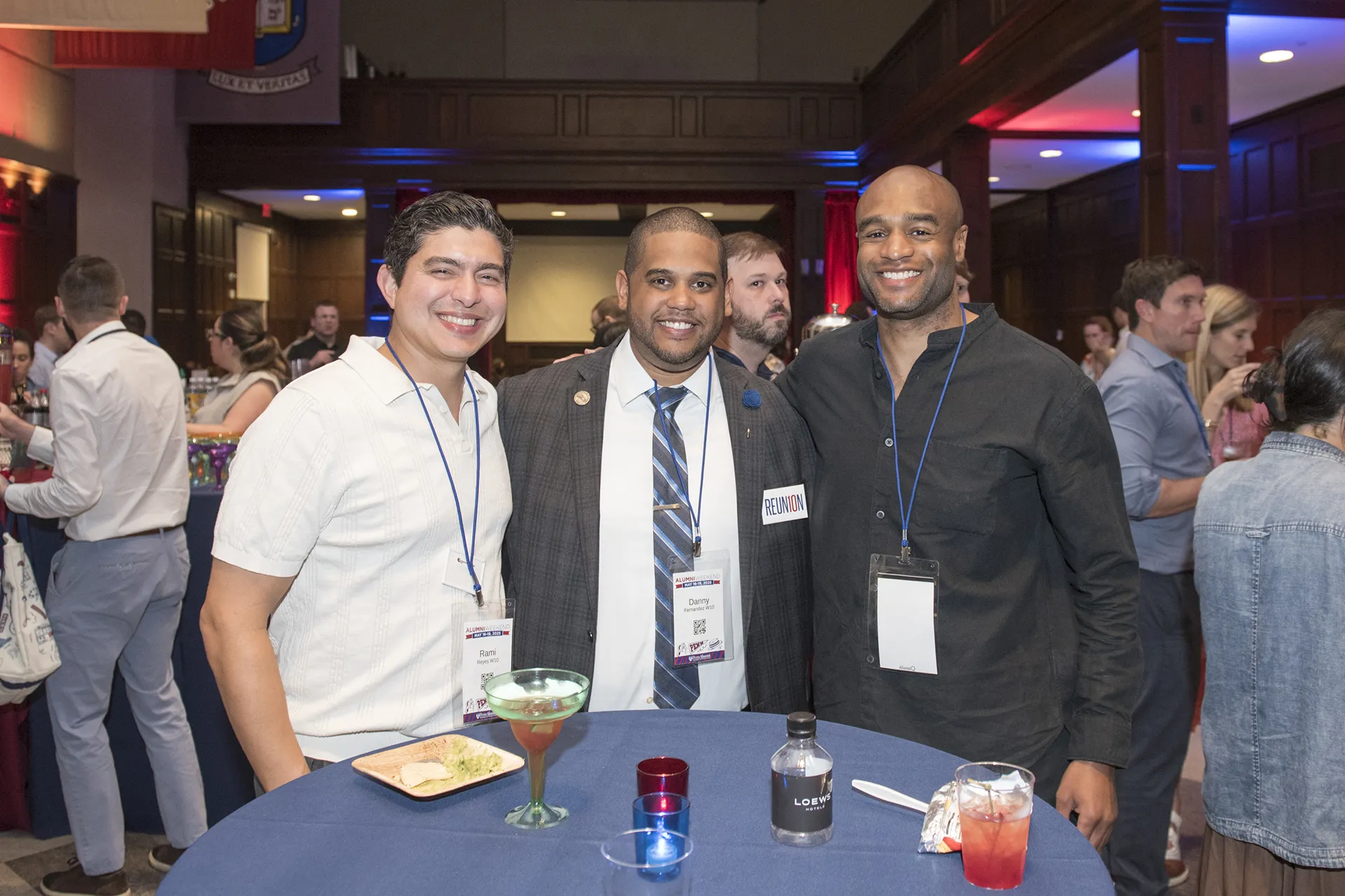 Three Penn alumni from the Class of 2010 smile together at their 15th Reunion reception, standing around a high-top table with cocktails and name badges